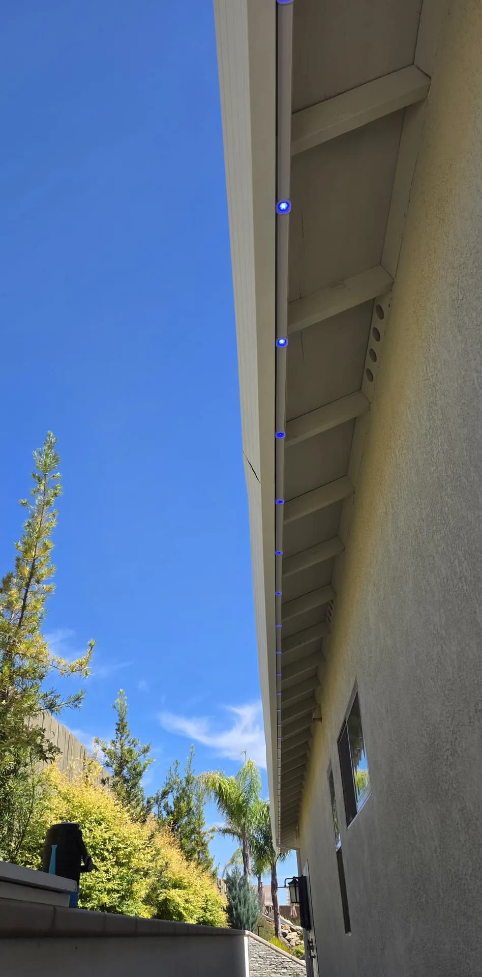 Permanent LED holiday lights along a fascia under open eaves, daytime